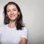 Portrait of a confident woman smiling with arms crossed, wearing a white t-shirt on a gray background.